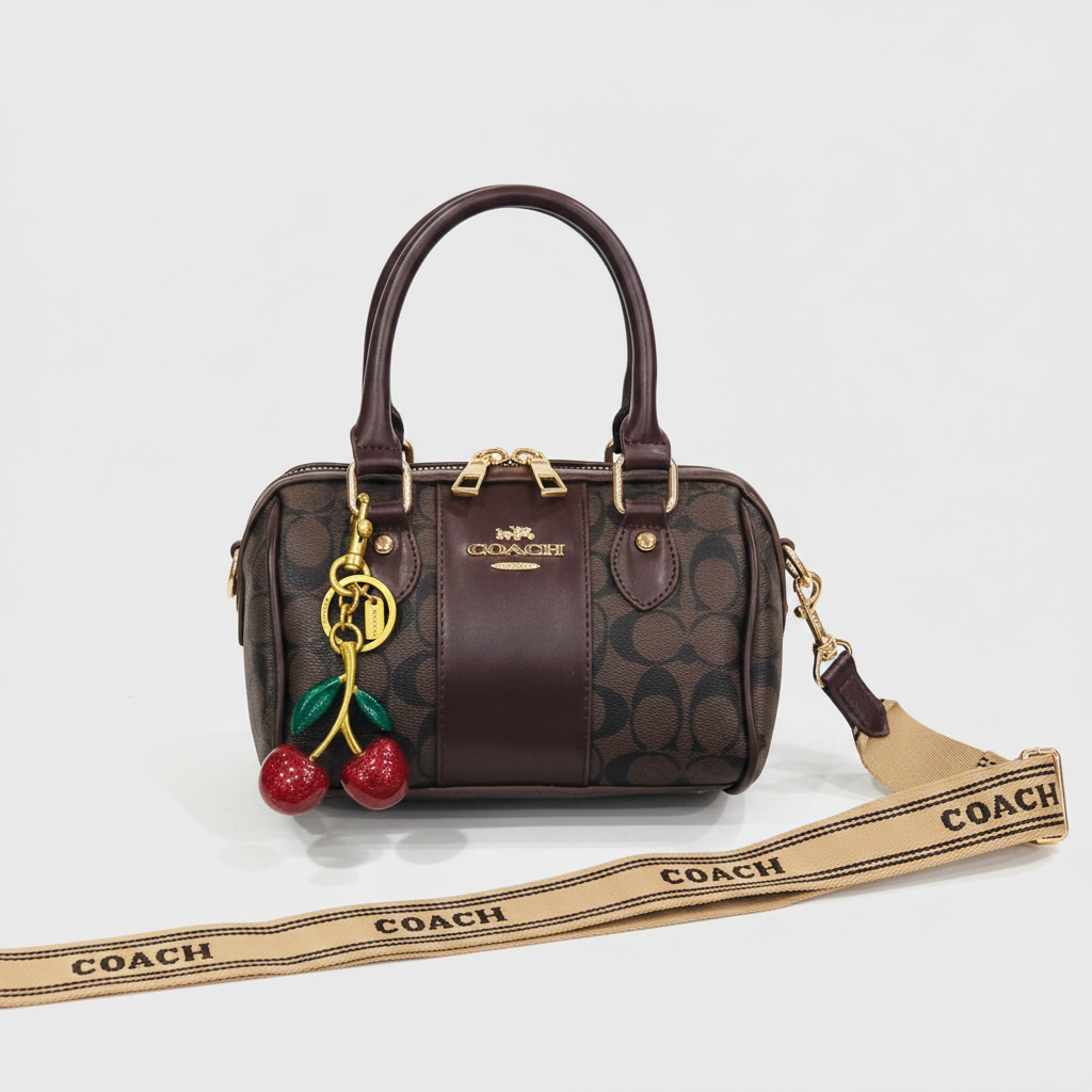 A Coach handbag in dark brown and beige monogram canvas with gold-tone hardware, featuring a cherry charm and a matching Coach shoulder strap laid out in front on a white surface.