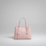 A light pink textured leather handbag with double top handles and a front strap detail, photographed against a neutral light gray studio background.