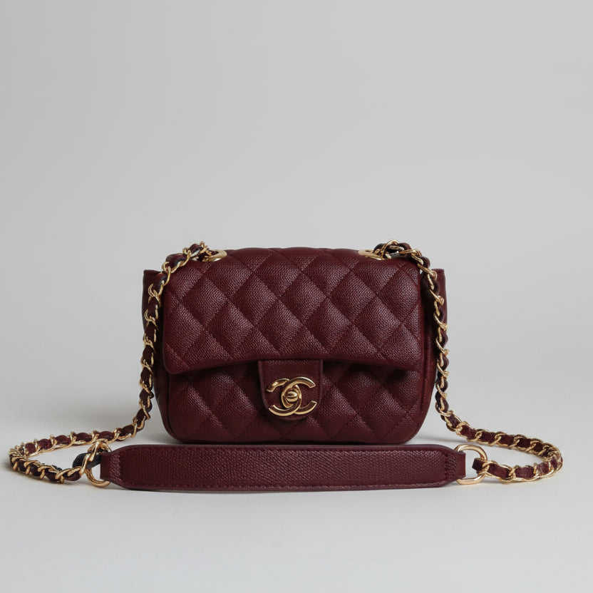 Maroon quilted handbag with gold chain strap on a light surface with a white textured wall background