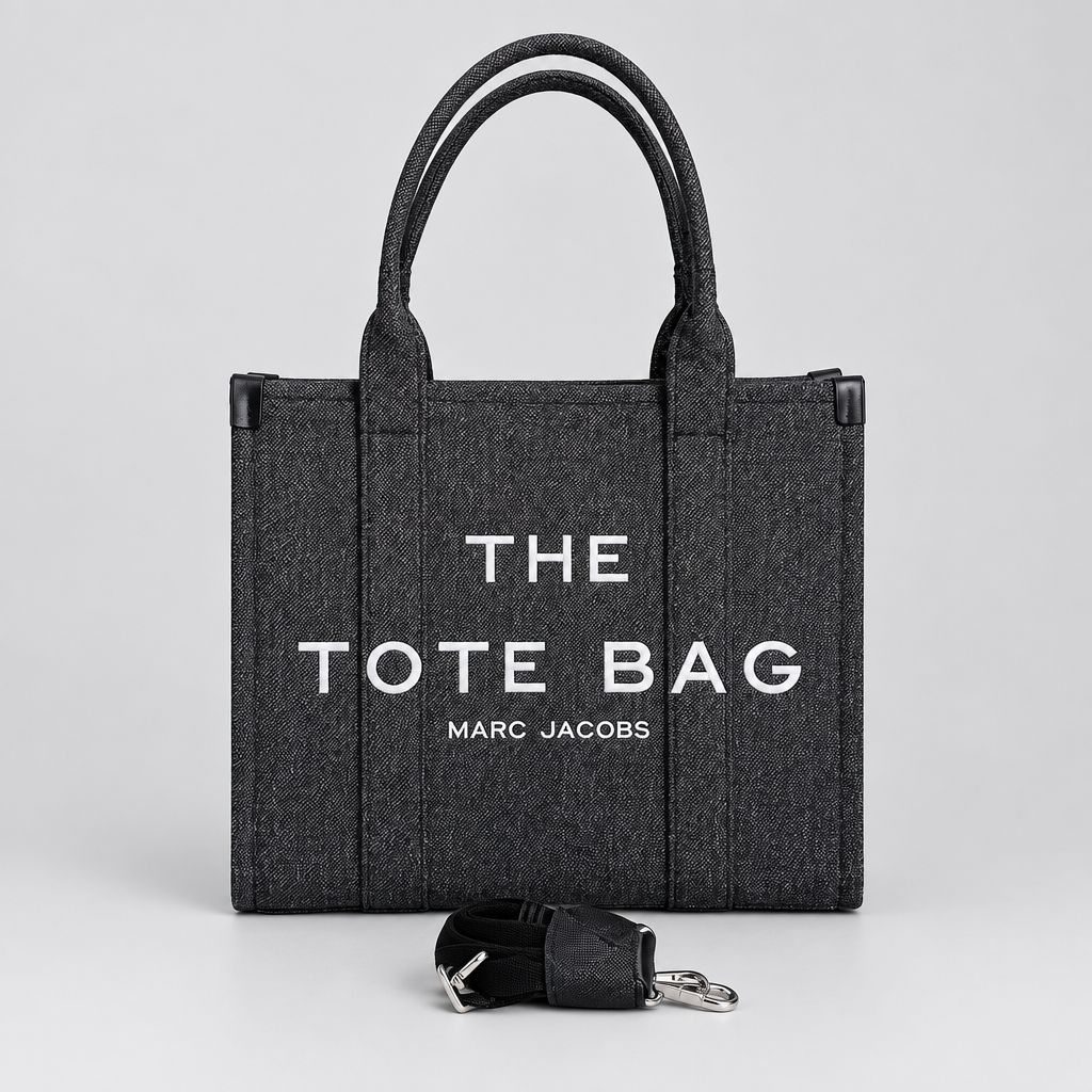 A dark gray woven tote bag with white text reading 'THE TOTE BAG' and 'MARC JACOBS' on the front, standing on a light surface against a white brick wall. A black detachable strap with a silver clasp lies in front of the bag.