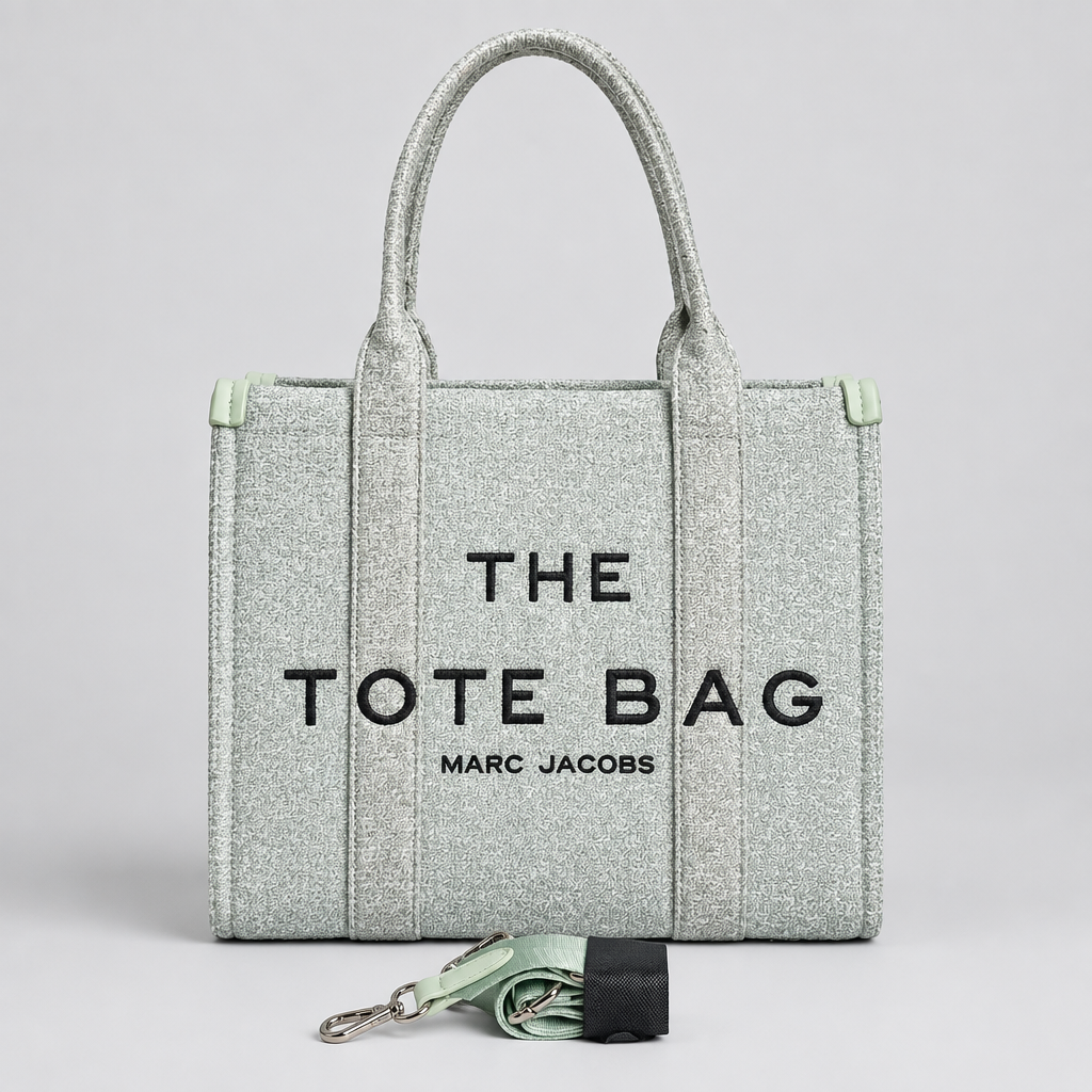 A light sage green canvas tote bag with black text reading 'THE TOTE BAG' and 'MARC JACOBS' on the front, standing upright against a white brick wall. The bag has two top handles and a matching rolled strap with a silver clasp resting in front.