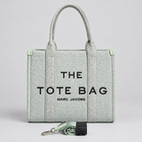 A light sage green canvas tote bag with black text reading 'THE TOTE BAG' and 'MARC JACOBS' on the front, standing upright against a white brick wall. The bag has two top handles and a matching rolled strap with a silver clasp resting in front.